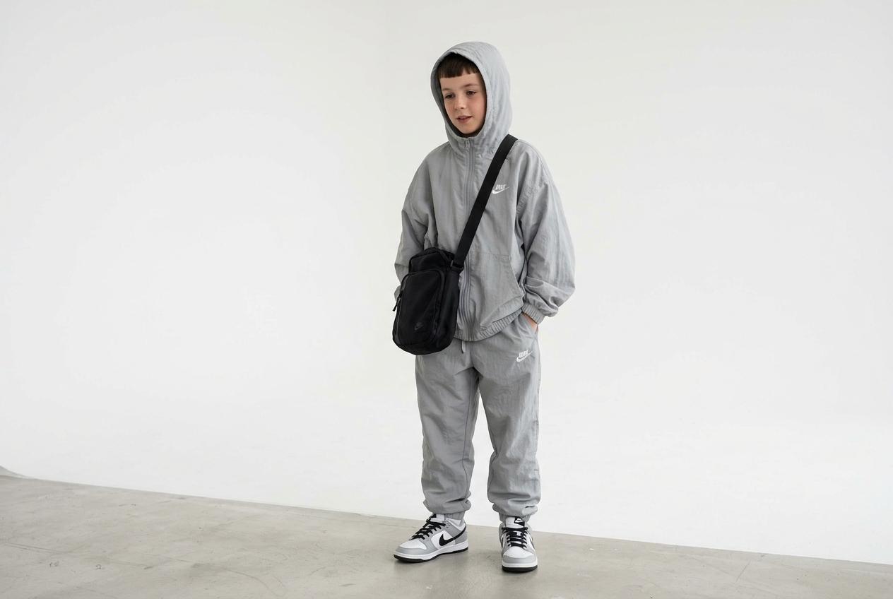 a young english 10-year old hooded teen boy with trim hair loitering with hands tucked into pockets dressed with crossbag close beside, over flowing, Wind tousled light grey hooded oversized zip up track suit with hood on, small white nike logo over joggers with white nike logo over nike dunk trainers in a white studio. CVY.AI
